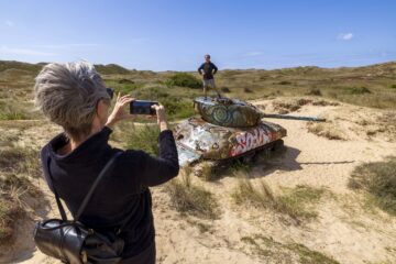 Char dans les dunes, un visiteur sur le toit du char et une personne le photographiant.