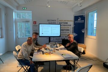 Trois hommes assis autour d'une table et dans le fond un écran tactile.
