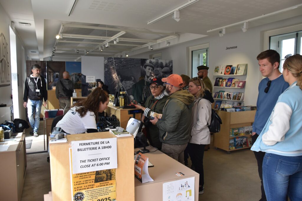 Accueil du site avec plusieurs visiteurs se rendant au comptoir de billetterie et de boutique, dans un espace lumineux proposant ouvrages, souvenirs et informations touristiques