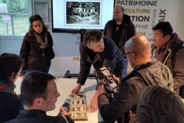 Plusieurs personnes (journalistes et équipe de la batterie d'Azeville) autour d'une table regardant l'album photo apporté par R. Graf