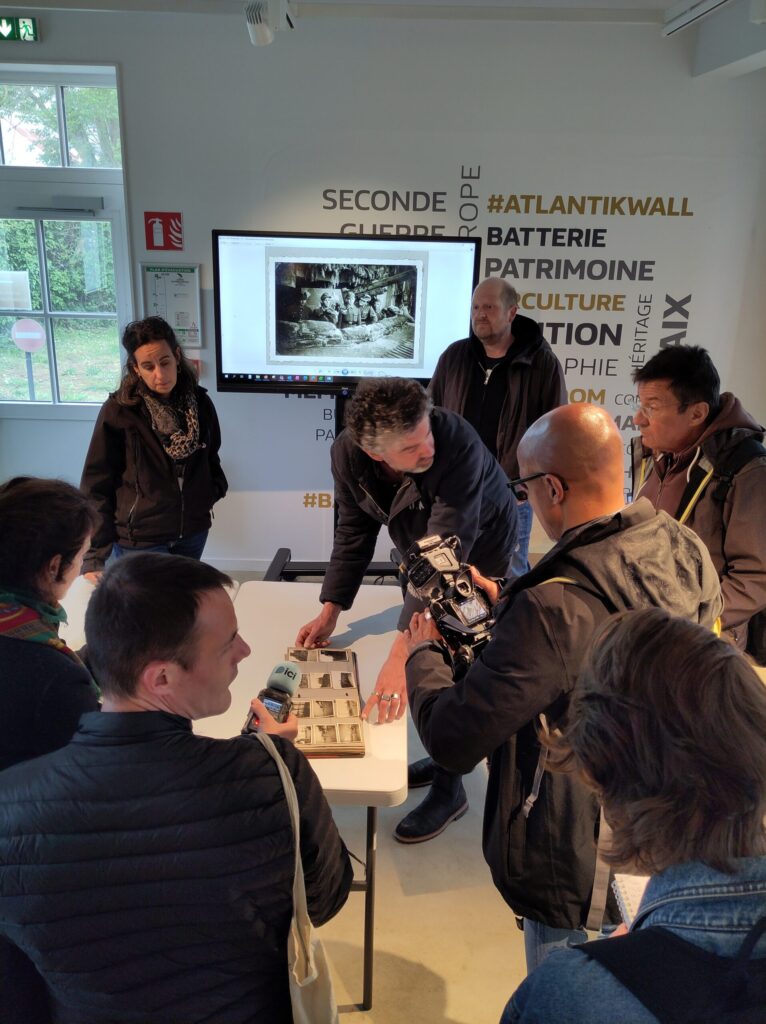 Plusieurs personnes (journalistes et équipe de la batterie d'Azeville) autour d'une table regardant l'album photo apporté par R. Graf